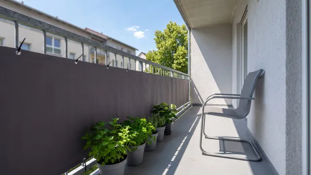 Symbolbild (KI-generiert): Balkon mit anthrazitfarbener Bespannung am Geländer, Kräutertöpfen und grauem Stuhl, Sommersonne