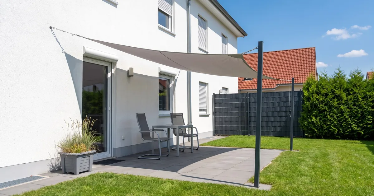 Symbolbild (KI-generiert): Terrasse mit hellgrauem Sonnensegel an Hauswand und zwei Pfosten, darunter Tisch und zwei Stühle im Schatten, Sommersonne
