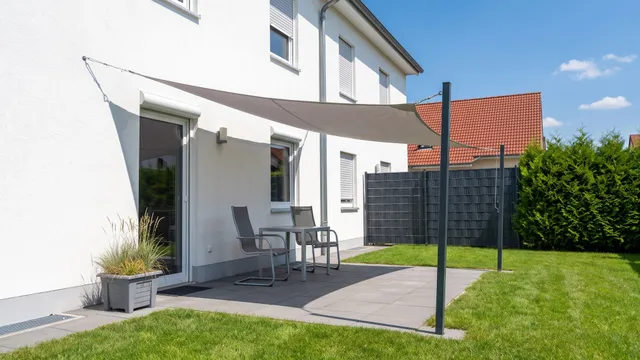 Symbolbild (KI-generiert): Terrasse mit hellgrauem Sonnensegel an Hauswand und zwei Pfosten, darunter Tisch und zwei Stühle im Schatten, Sommersonne